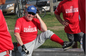 A smiling person wearing a red Falmouth Toyota t-shirt and blue cap rests on a bench in a grassy outdoor setting.