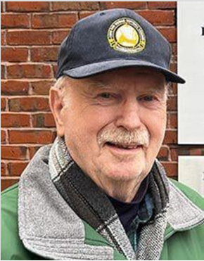 A smiling person wearing a dark baseball cap with a gold emblem, a grey scarf, and a green jacket against a brick wall.