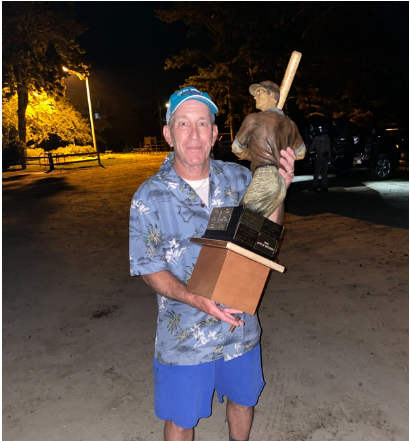 A person wearing a blue patterned shirt and hat holds a large baseball trophy at night in an outdoor parking lot.