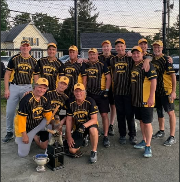 A team of softball players wearing matching dark shirts poses for a photo on a dirt field, holding a trophy and baseballs.
