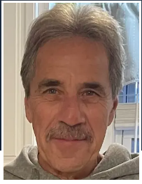 A portrait of a person with gray-brown hair and a mustache, wearing a gray hooded sweatshirt, looking toward the camera.