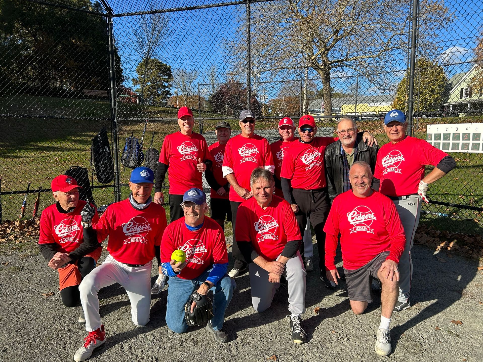 A group of people wearing matching red team shirts stand and kneel on a gravel field in front of a chain-link fence.