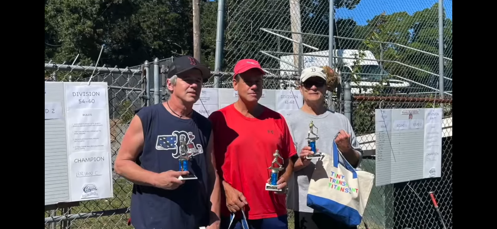Three people standing in front of a chain-link fence, holding small trophies, with score sheets attached to the fence.