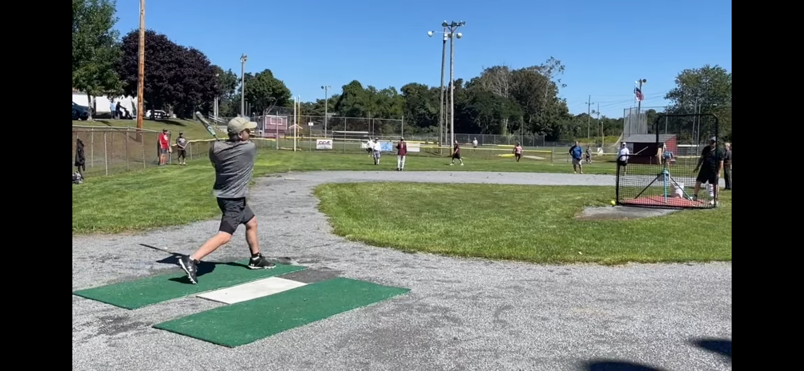 A person in a gray shirt and hat follows through with a baseball swing on an outdoor gravel practice field.