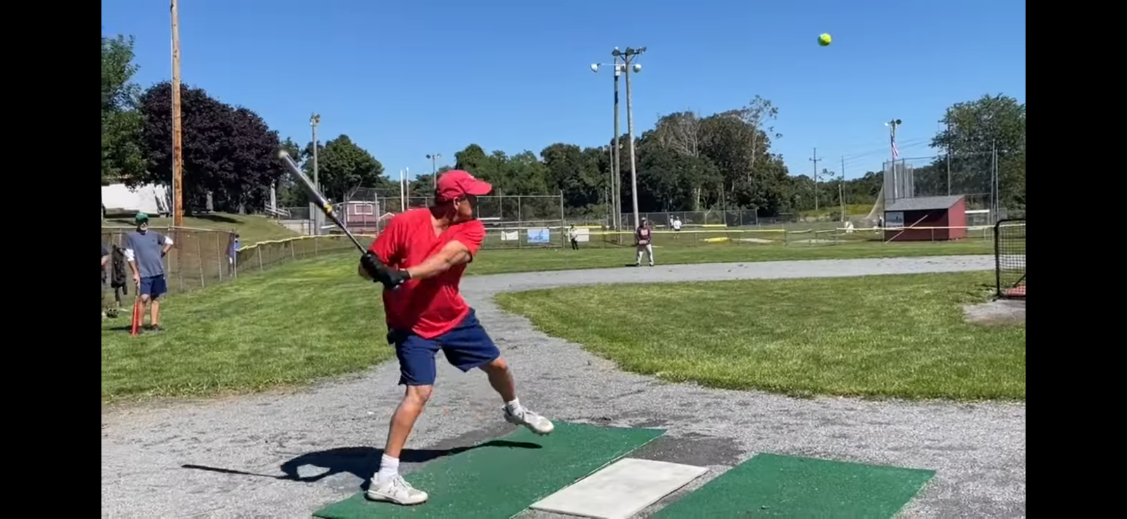 A person in a red shirt and blue shorts swings a bat at a softball in an outdoor batting cage on a sunny day.