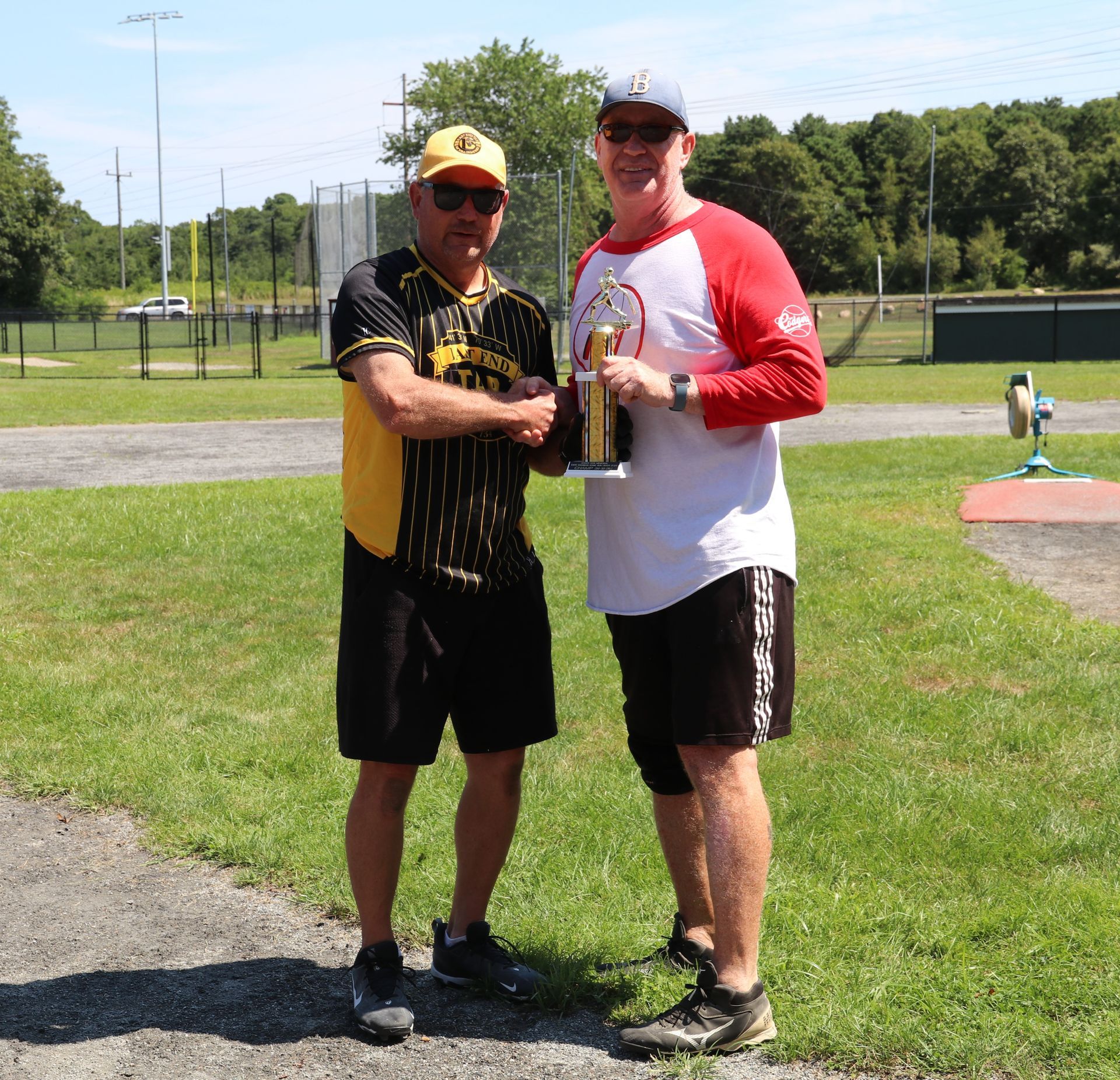 Two people shaking hands on a baseball field, one holding a trophy while wearing a white and red jersey.