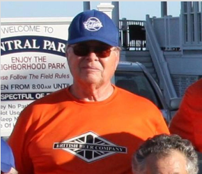 A person wearing a blue cap and orange British Beer Company t-shirt stands in front of a neighborhood park sign.