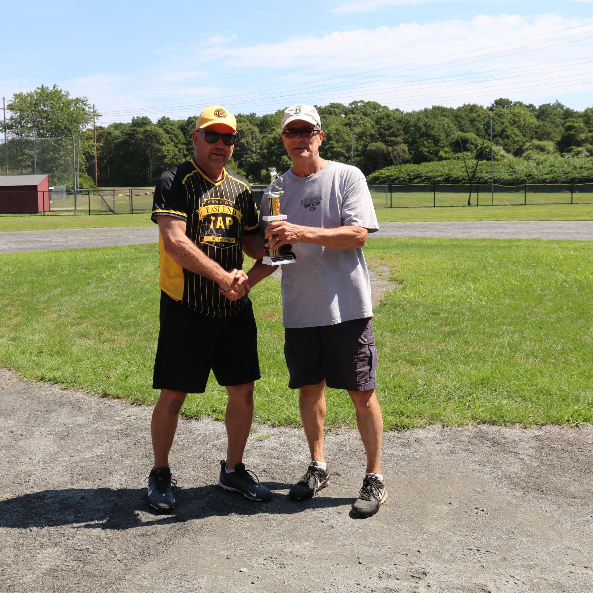 Two people stand on a grassy field, shaking hands while holding a trophy, wearing casual athletic clothing and hats.
