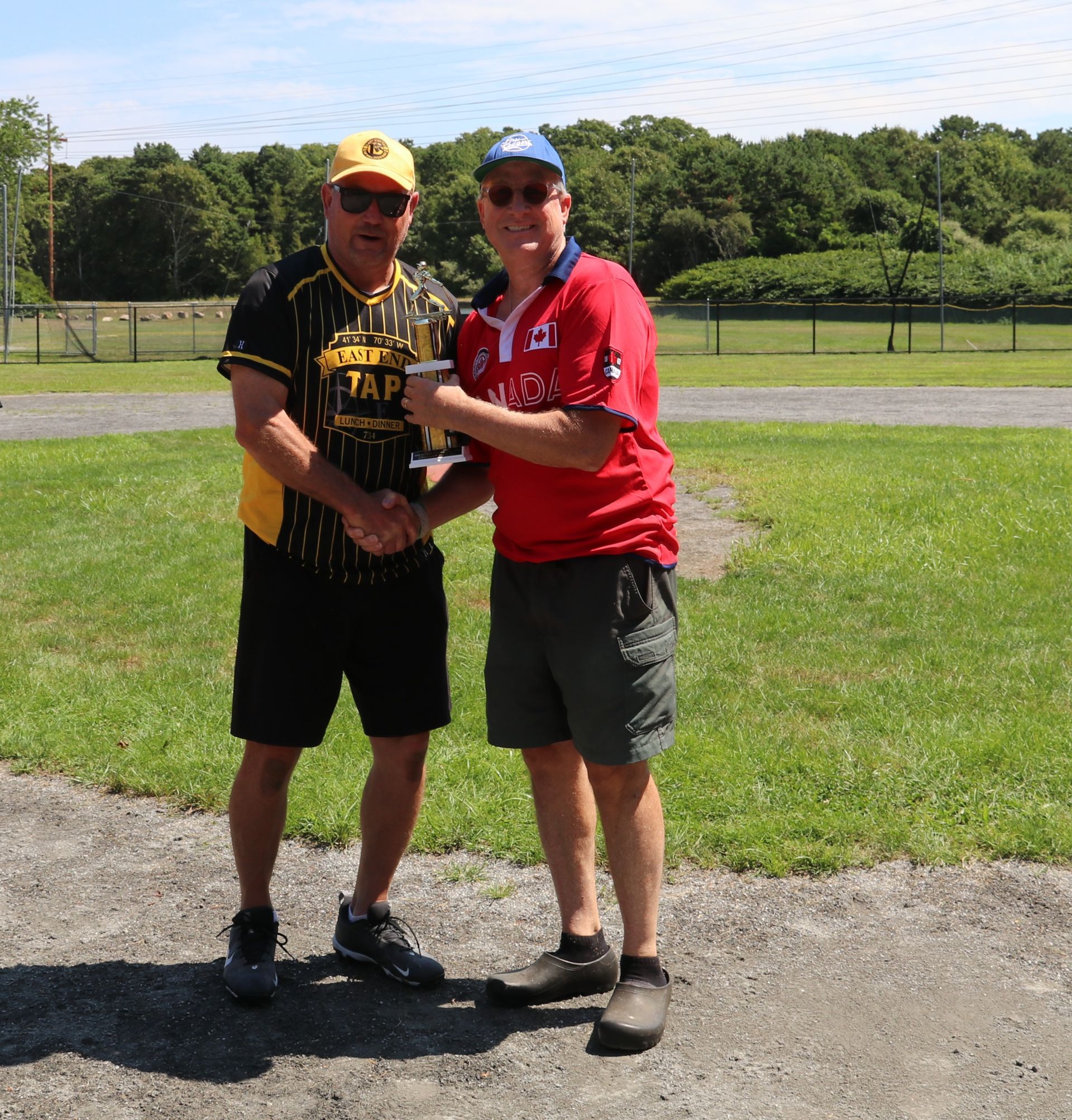 Two individuals stand outdoors on a field, shaking hands while holding a small trophy, both wearing hats and sunglasses.