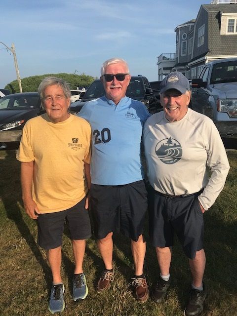 Three individuals smiling and posing outdoors on a sunny day, wearing casual shirts and shorts near parked vehicles.