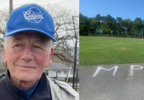 An older man in a blue baseball cap next to a grassy field with the letters MP spray-painted on the pavement.