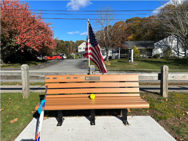 A tan bench with a memorial plaque sits on a concrete pad, adorned with an American flag, a baseball bat, and a yellow ball.