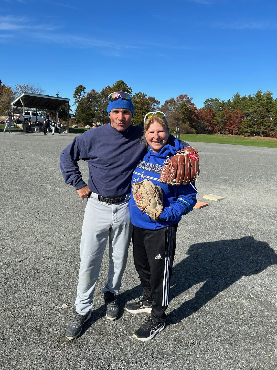 Two people pose on a gravel field, one holding a baseball glove, under a clear blue sky.