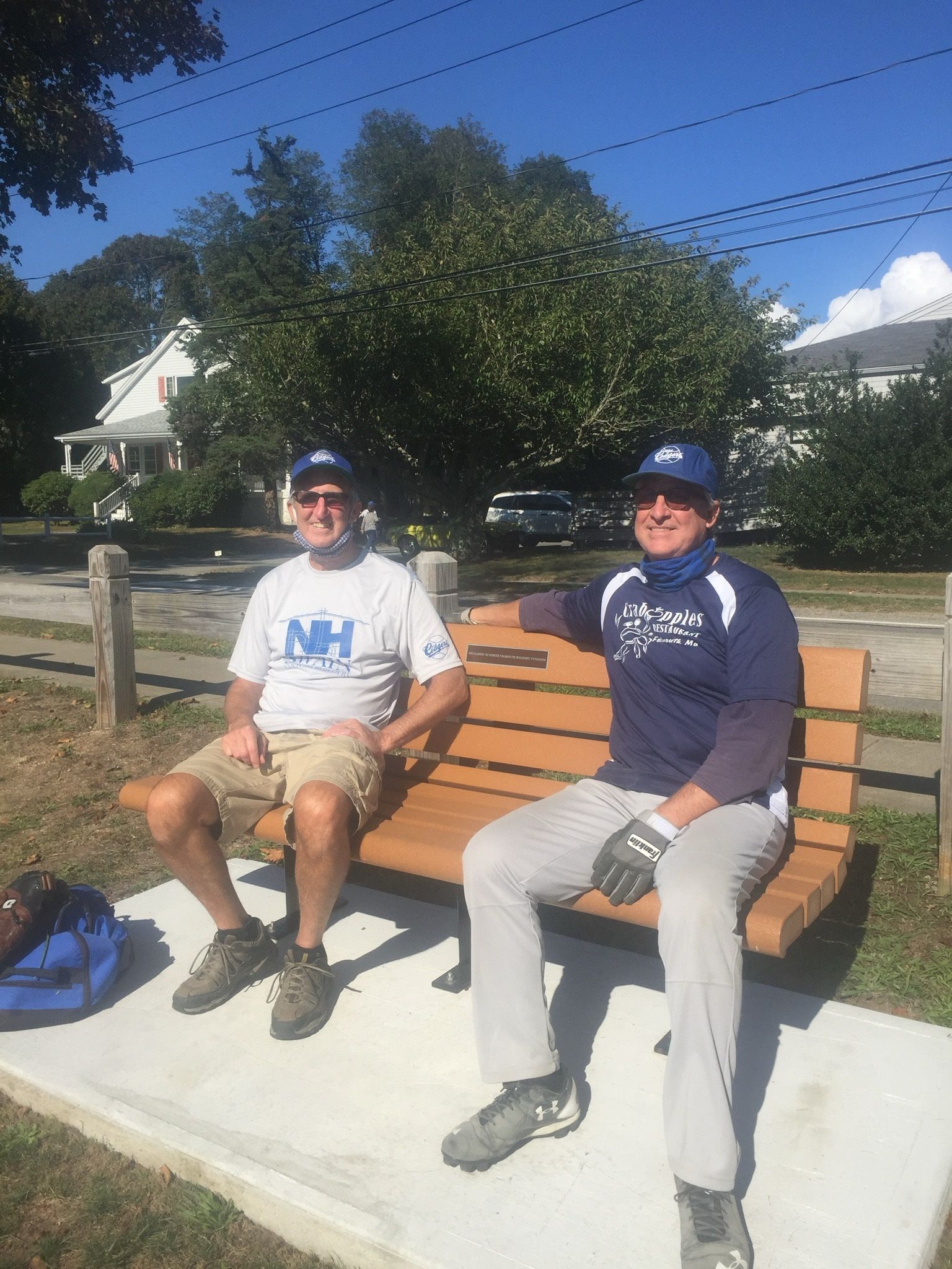 Two people in caps sit on a tan bench on a concrete pad outdoors on a sunny day.