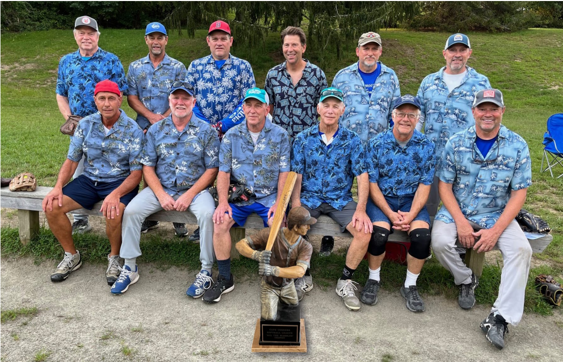 A baseball team in blue floral shirts poses outdoors on a bench with a trophy featuring a batter figure.
