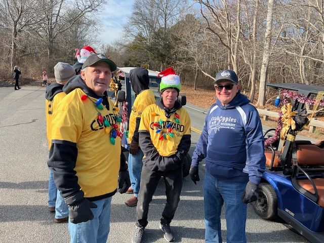 Three people in casual winter attire and holiday hats, two wearing festive yellow shirts, standing by a blue golf cart.