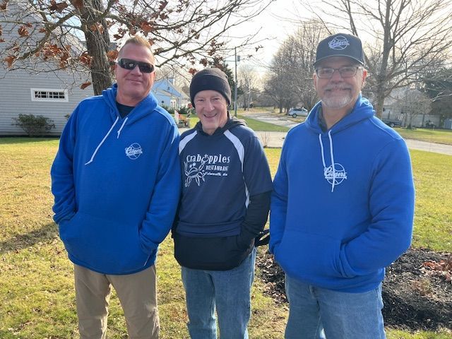 Three people standing outdoors in a yard, wearing casual clothing and matching blue hooded sweatshirts with a logo.