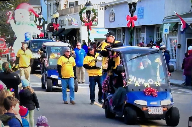 People in yellow shirts walk beside two blue golf carts decorated with festive red bows in a small-town holiday parade.
