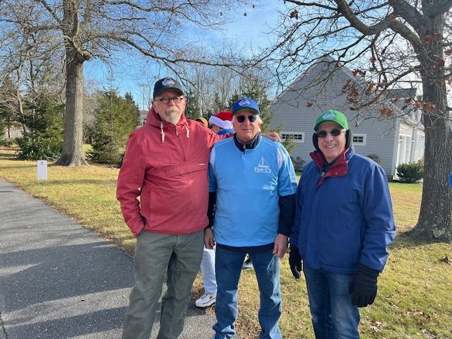 Three people wearing hats and jackets stand outside on a sunny day in front of a house and trees.