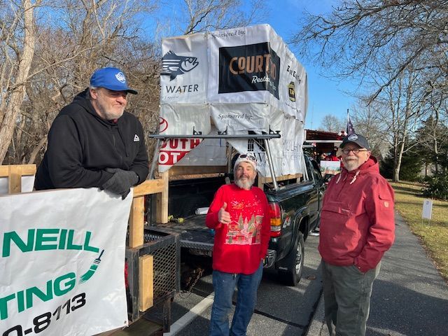 Three people stand by a truck loaded with cargo on a sunny day. A banner for O’Neill Roofing is visible in the foreground.