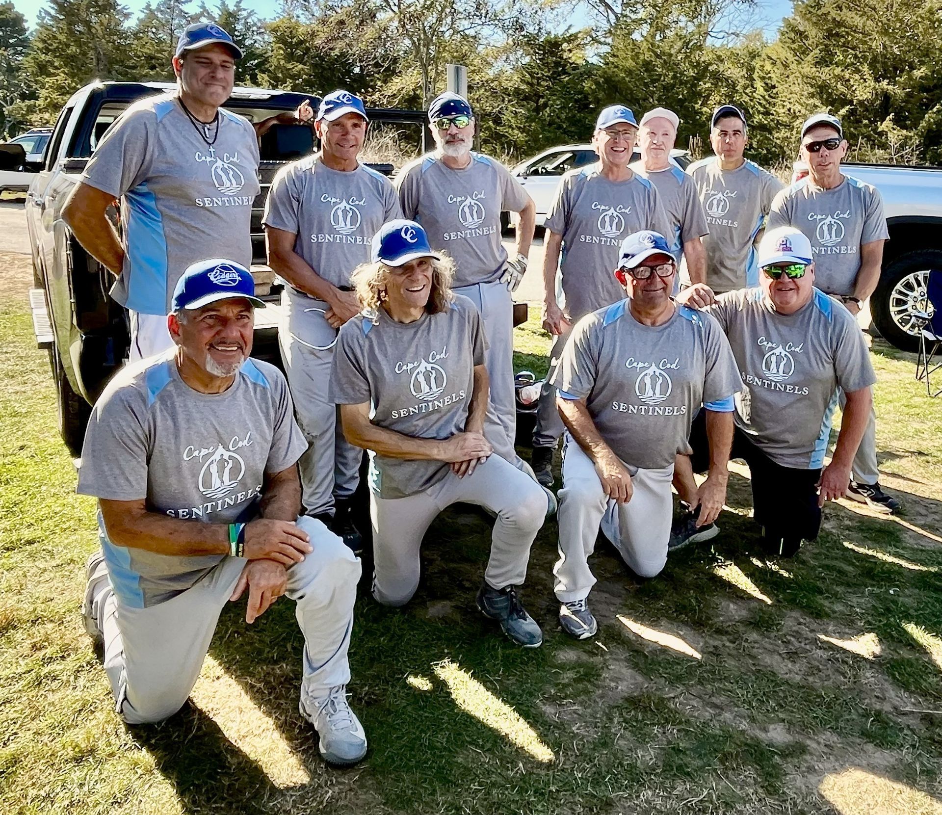 A group of men in matching grey t-shirts and baseball caps pose outdoors in a sunny, grassy field.