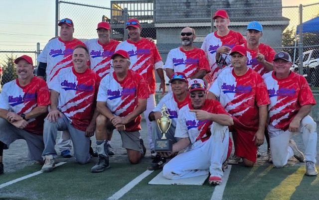A softball team in red and white jerseys poses for a group photo holding a gold trophy on an outdoor field.