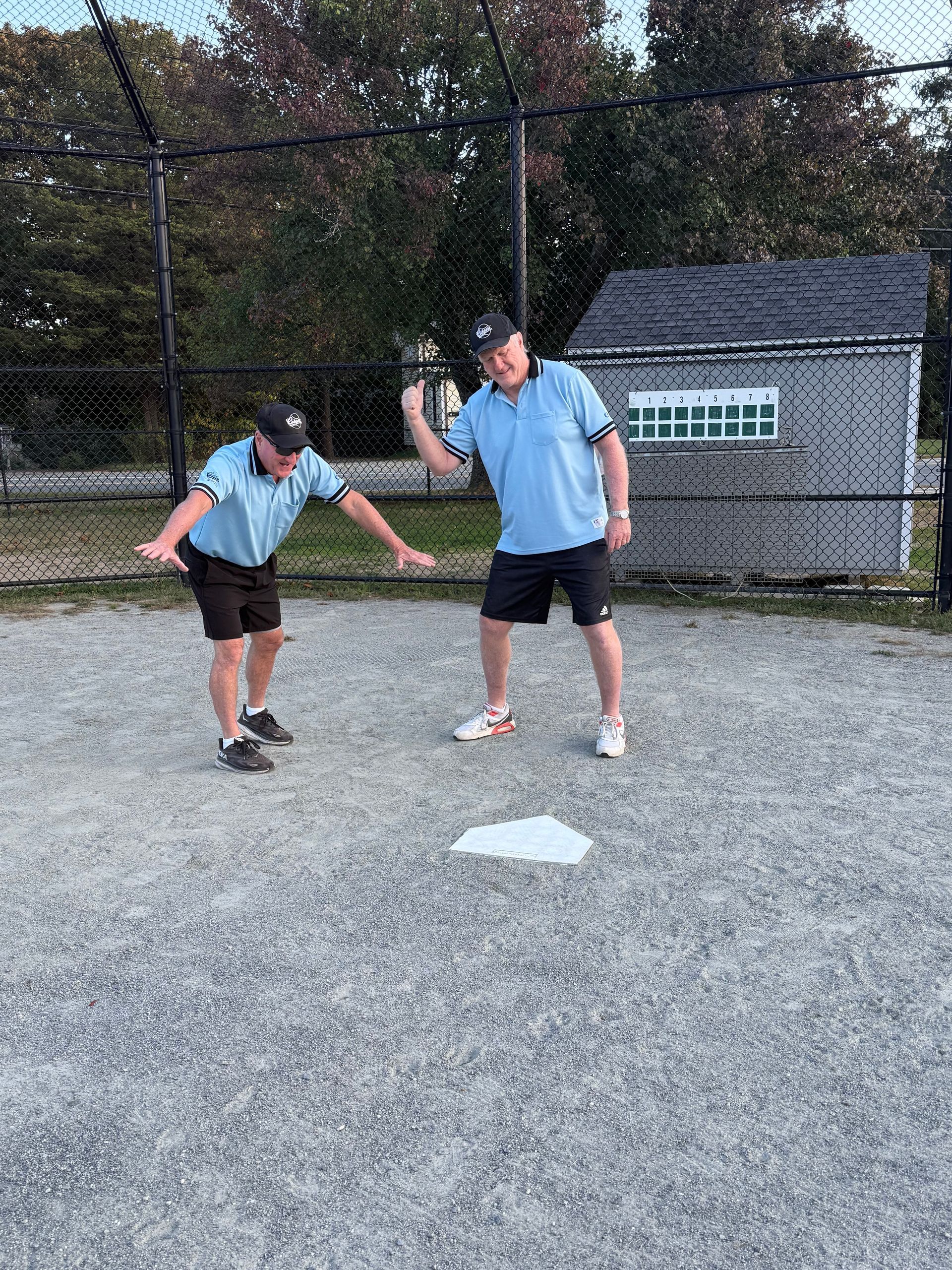 Two people in light blue shirts and dark shorts pose on a baseball field near home plate.