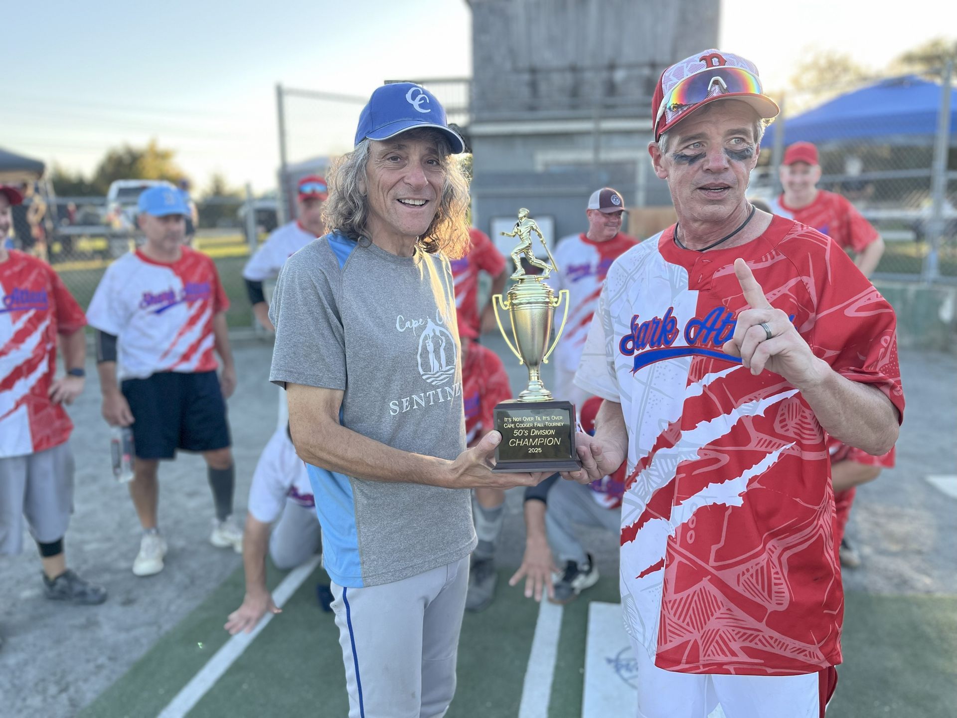 Two individuals hold a trophy on a softball field, flanked by teammates in matching red and white sports uniforms.