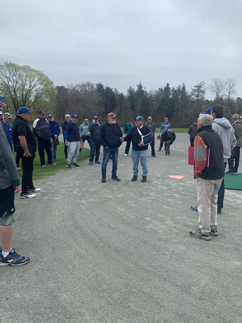 A group of people wearing casual clothing stand in a circle on a gravel baseball field during an outdoor gathering.