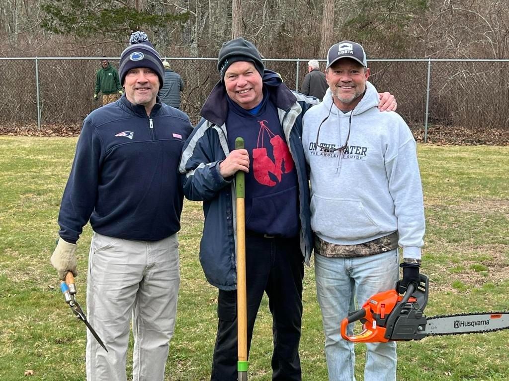 Three people standing on a grassy field, holding gardening tools and a chainsaw, posing together in a park setting.
