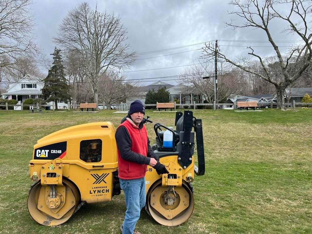 A person wearing a red vest and blue beanie stands next to a yellow CAT steamroller in a grassy park.