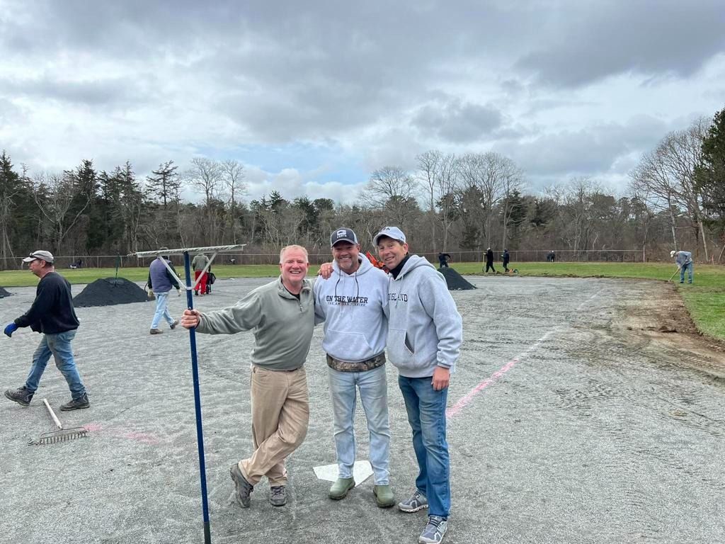 Three people pose on a baseball field under construction, with one holding a landscape rake, under a cloudy sky.