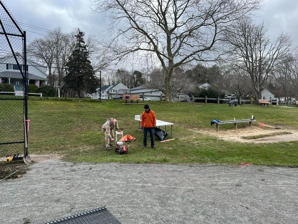 Two people work with equipment on a grassy field near a gravel area, a fence, and trees under a cloudy sky.