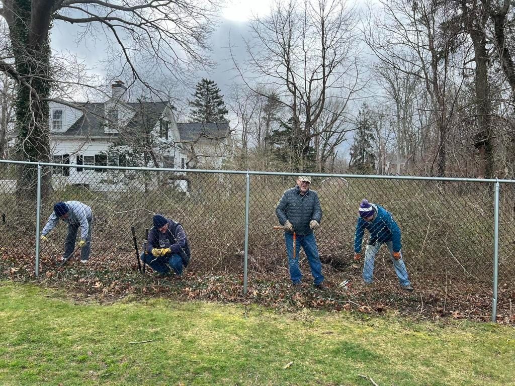Four people working together to clear brush and yard debris along a chain-link fence in front of a house.