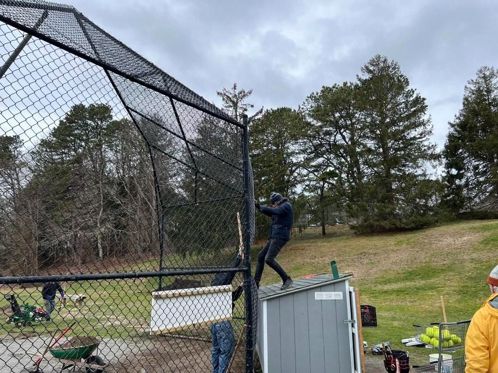 A person stands on top of a gray shed, reaching up to repair or install netting on a chain-link fence at a sports field.