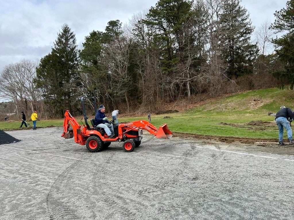 An orange tractor moves across a gravel lot near a grassy field and wooded area, with people working nearby.