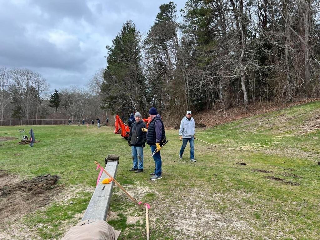Three people standing on a grassy field near a forest edge, observing construction equipment and a marked layout line.