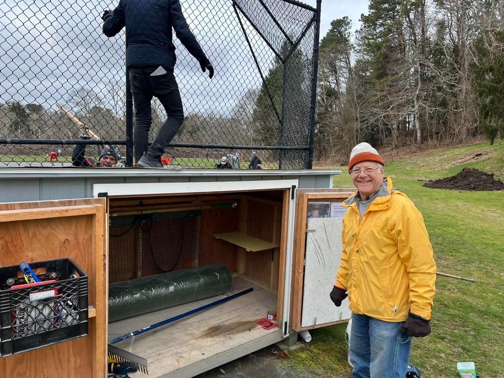 A person in a yellow jacket stands by an open outdoor storage unit, while someone stands on the roof above it.