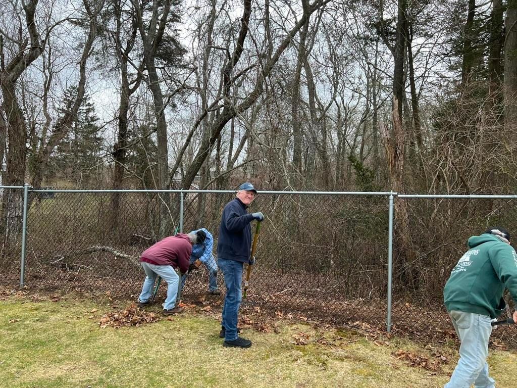 Three people in outdoor clothing work along a chain-link fence, clearing brush and leaves near a wooded area.