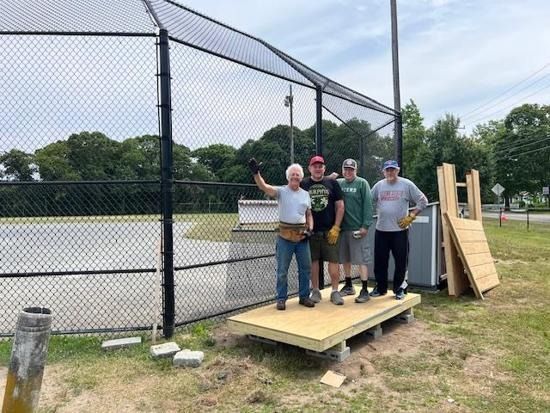 Four people stand on a newly built wooden platform in front of a baseball field fence, smiling and posing for a photo.