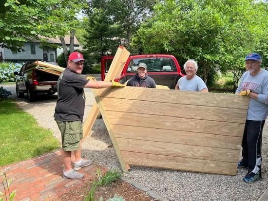 Four people standing in a driveway, holding a large wooden structure outdoors near a pickup truck.
