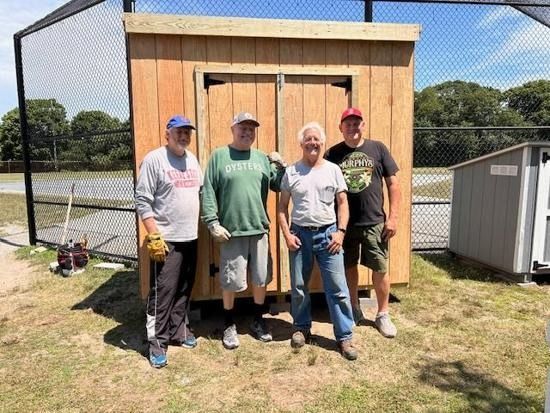 Four smiling people stand side-by-side in front of a newly constructed wooden shed at a baseball field.