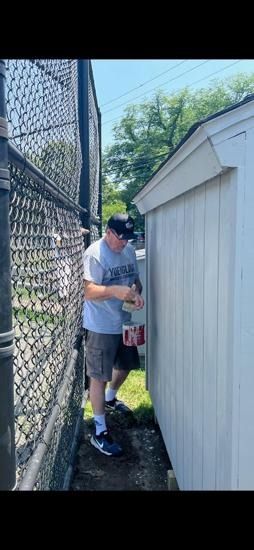 A person wearing a t-shirt, shorts, and a cap paints the exterior wall of a shed next to a chain-link fence.