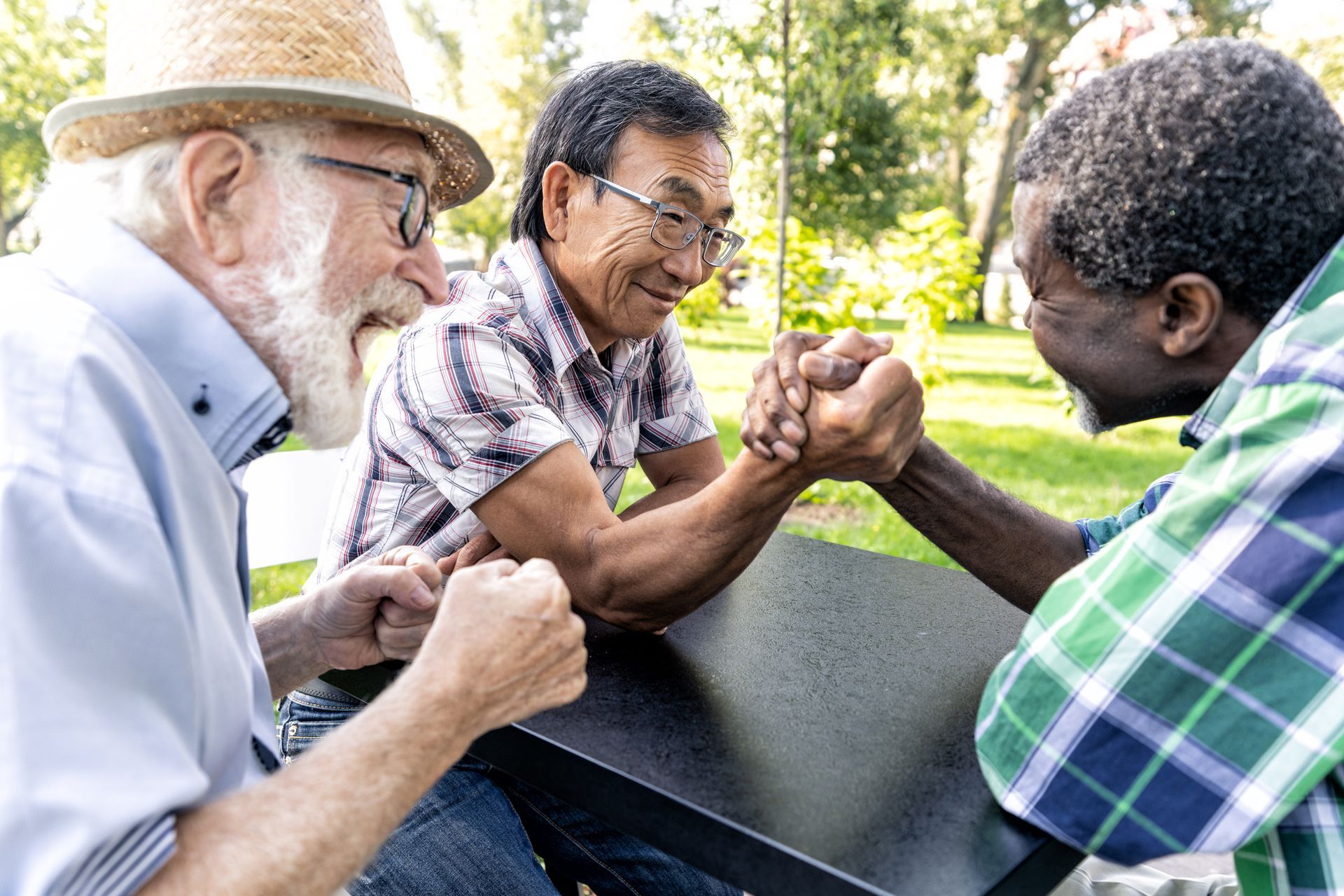 Three people sitting at an outdoor table, smiling as two of them engage in a friendly arm-wrestling match.