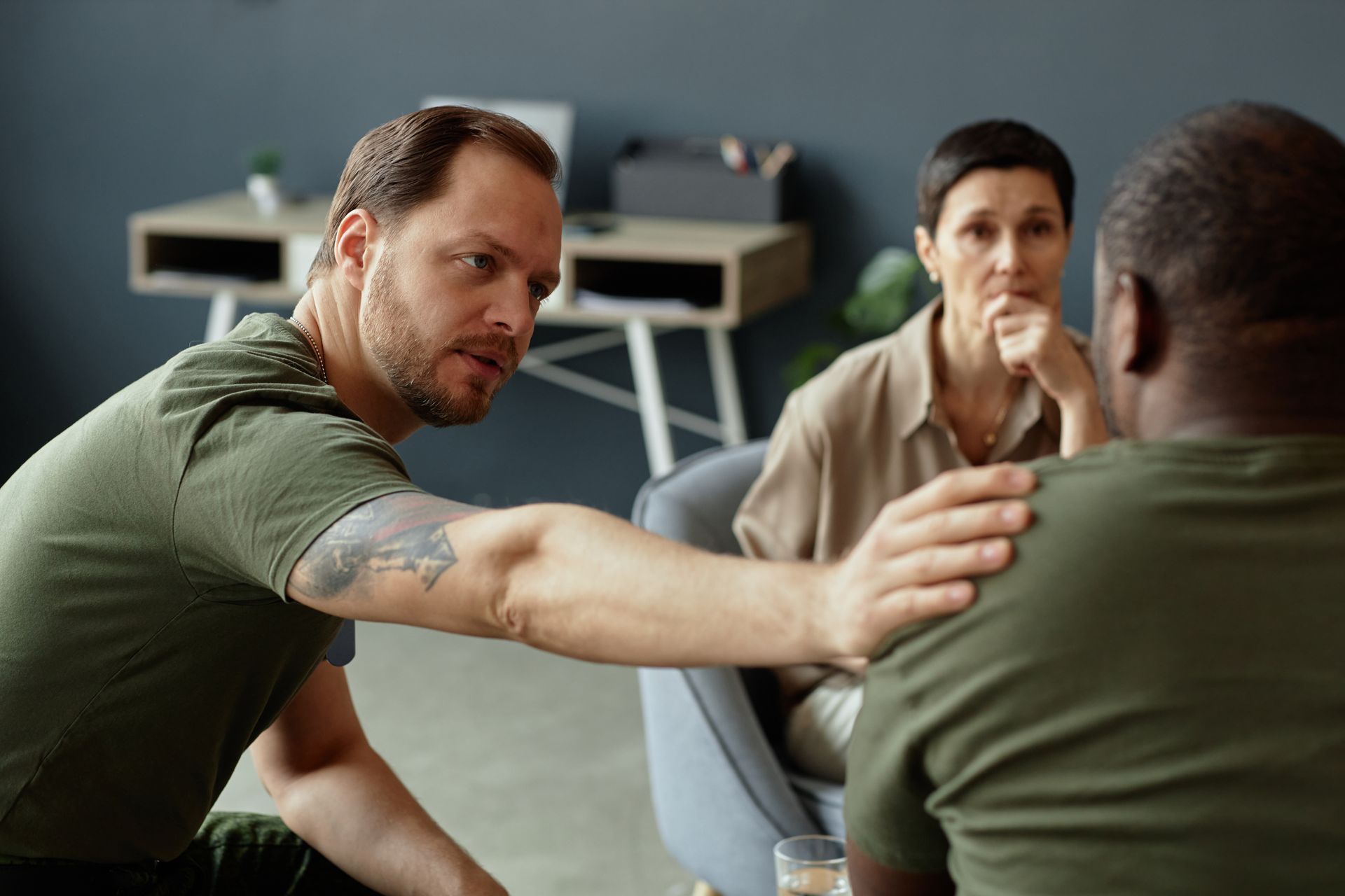 A man in a green t-shirt offers comfort by placing a hand on another person's shoulder while a third person looks on.