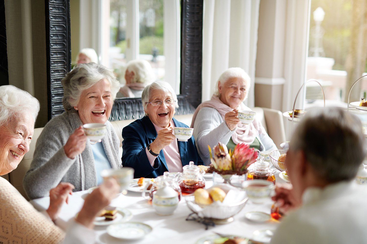Group of older adults laughing and toasting at a bright dining table with flowers and tea cups