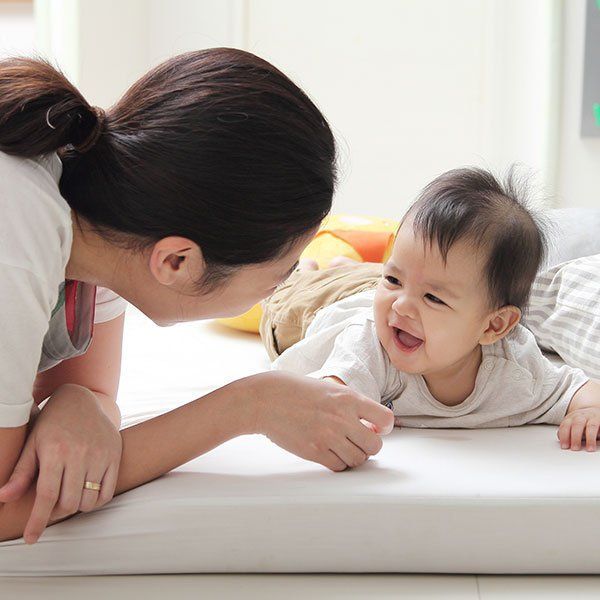 Woman leaning toward smiling baby on a white bed, playing face-to-face in a bright room