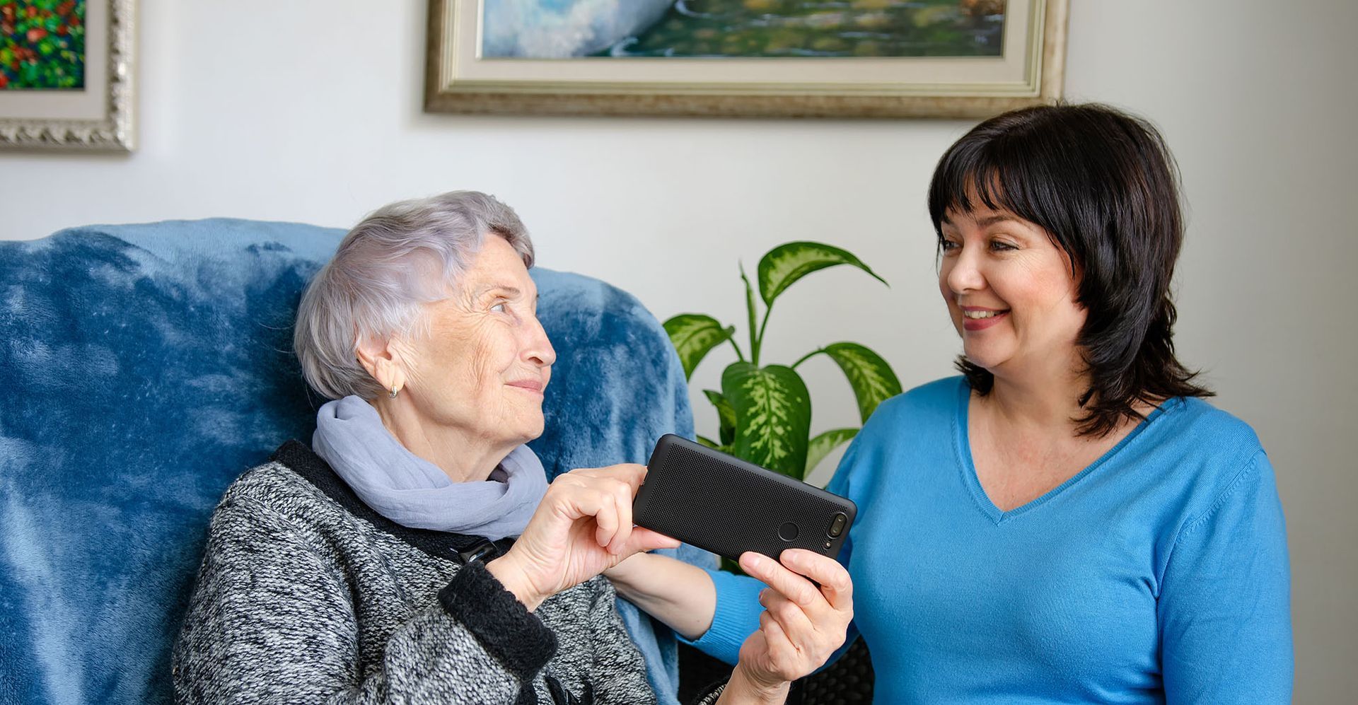 Older person and woman smiling while looking at a smartphone in a cozy living room.
