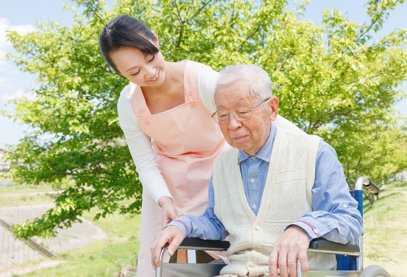 A caregiver helps an elderly man in a wheelchair outdoors under bright green trees.
