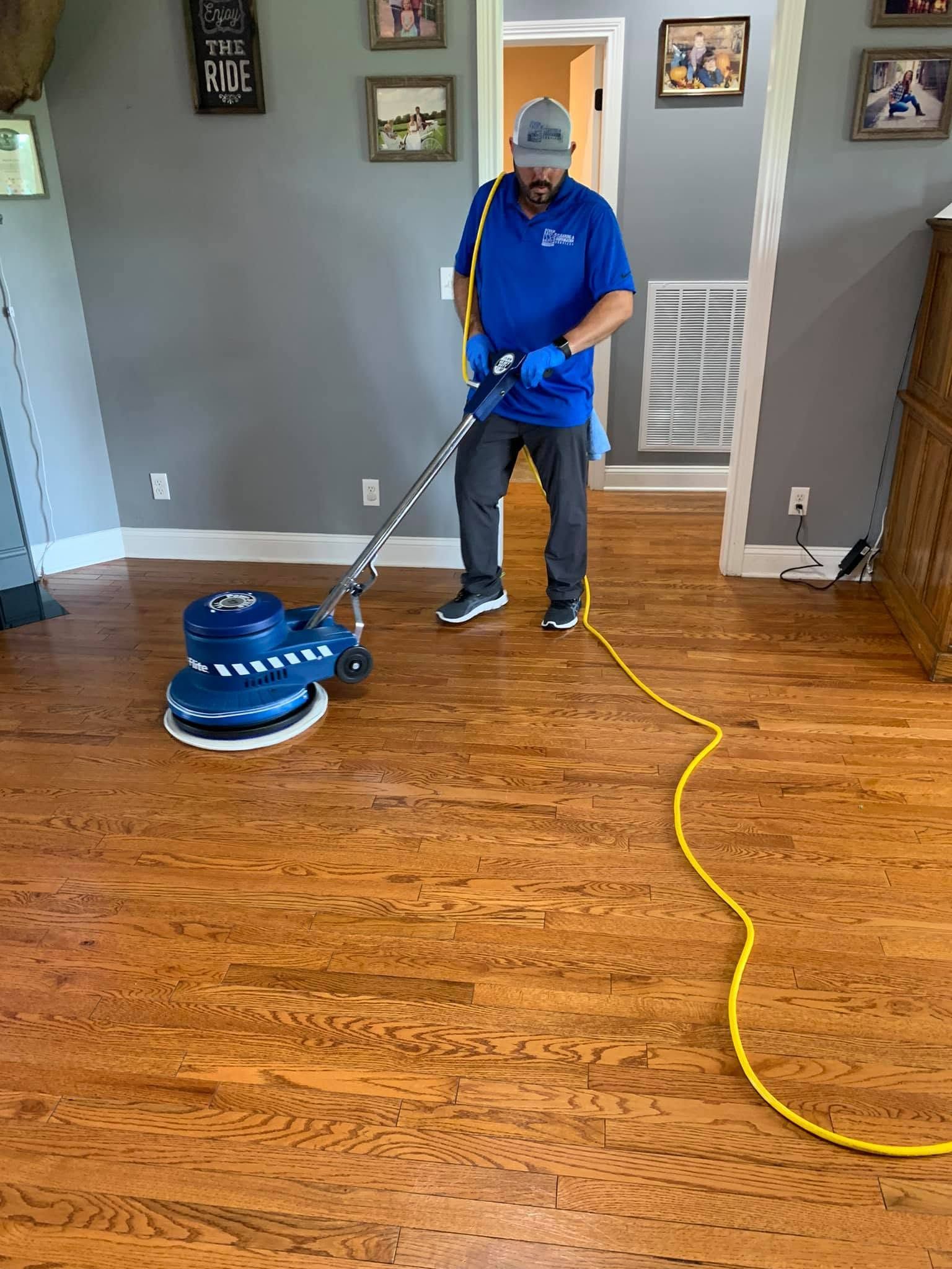 Person using a blue floor buffer on a hardwood floor in a room, with a yellow extension cord.